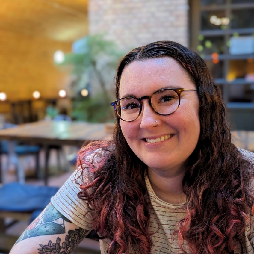 picture of a smiling woman, wearing glasses. she has long curly hair, dyed red, and tattoos on her arm, including a cactus.